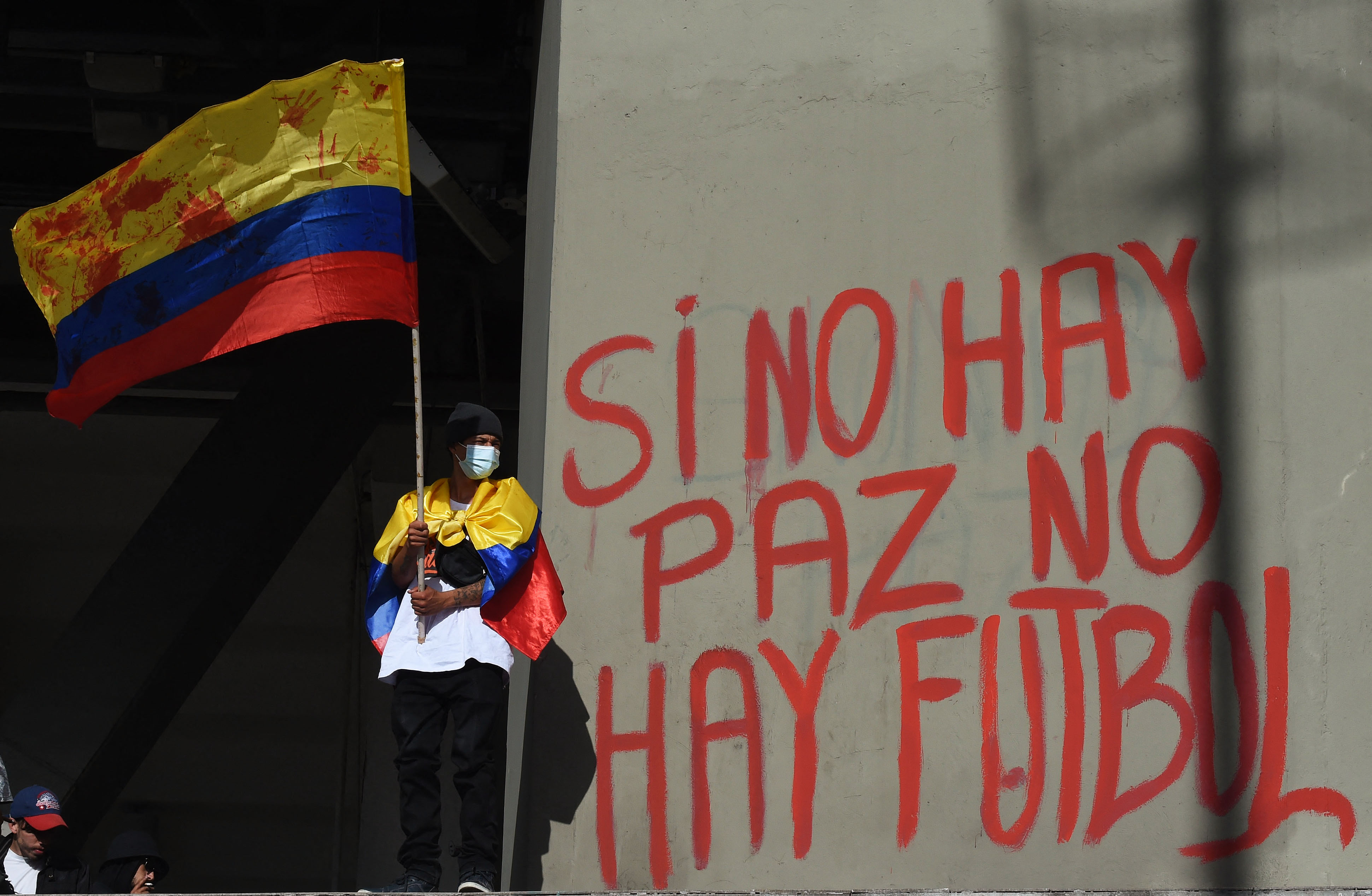 Un manifestante sostiene una bandera nacional colombiana junto a un graffiti en una pared del estadio El Camp&iacute;n que dice 'No hay paz, no hay f&uacute;tbol' durante una protesta contra Colombia como sede del torneo de f&uacute;tbol Copa Am&eacute;rica frente al estadio El Camp&iacute;n en Bogot&aacute;, el 19 de mayo de 2021 mientras el pa&iacute;s enfrenta su peor crisis pol&iacute;tica y civil en la historia reciente.